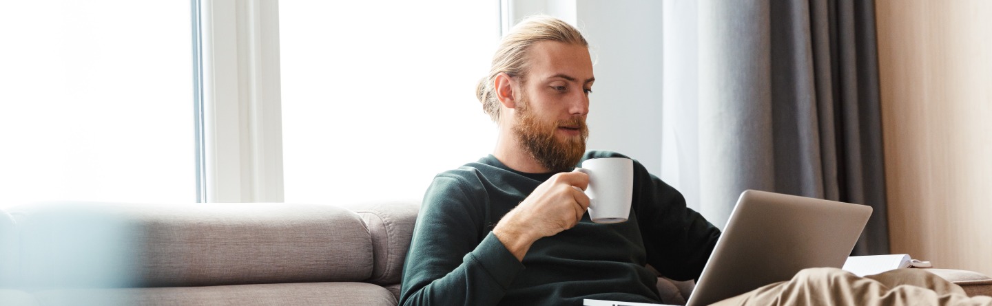 Ein junger Mann sitzt mit einem Laptop und einer Tasse auf einem Sofa